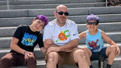 Ryan Roy sitting on outdoor steps between his two sons at a Walk MS event, all three smiling in bright sunshine